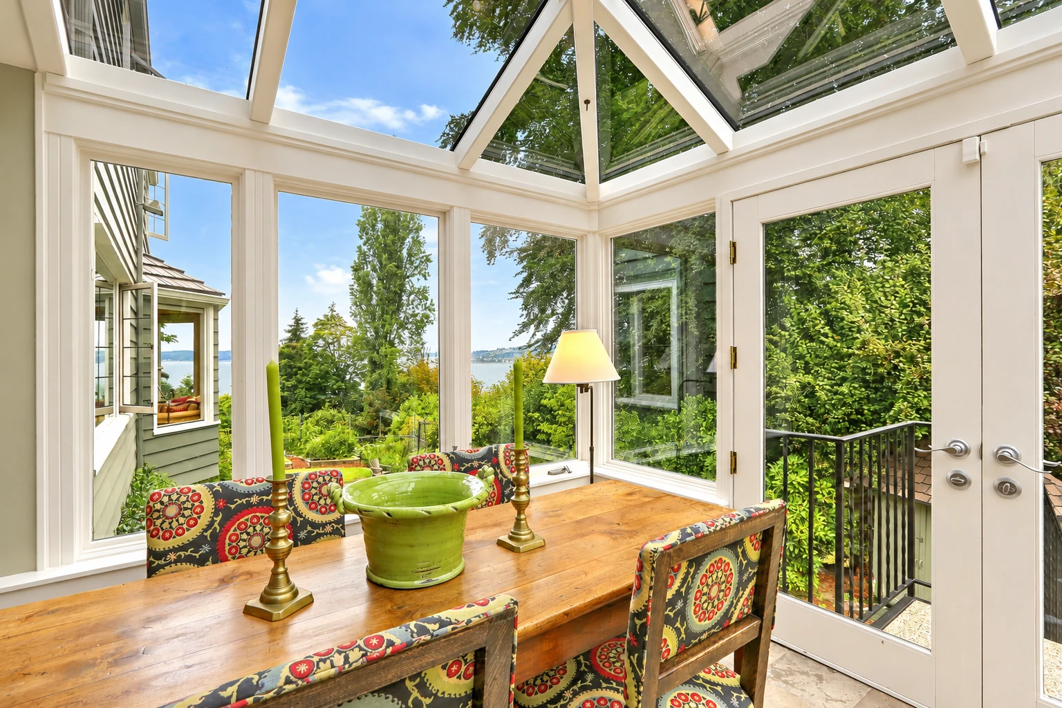 Custom sunroom with a glass roof and large windows, featuring a wooden dining table and colorful chairs among leafy views.
