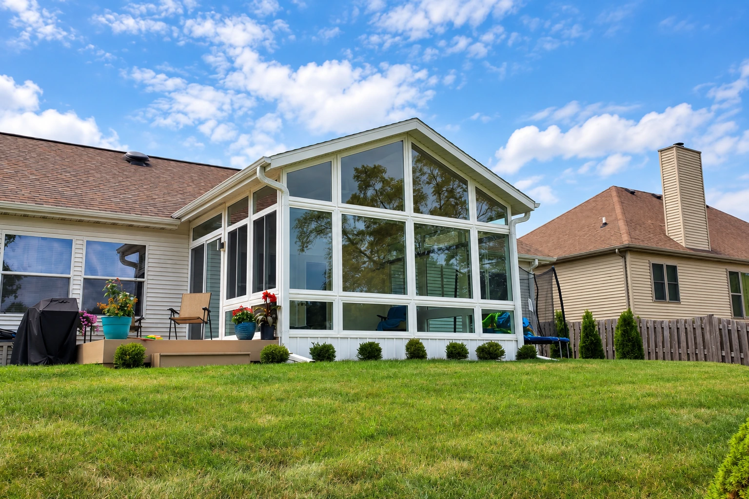 White-framed, gabled glass sunroom extends from a house above a lawn, with patio furniture nearby.