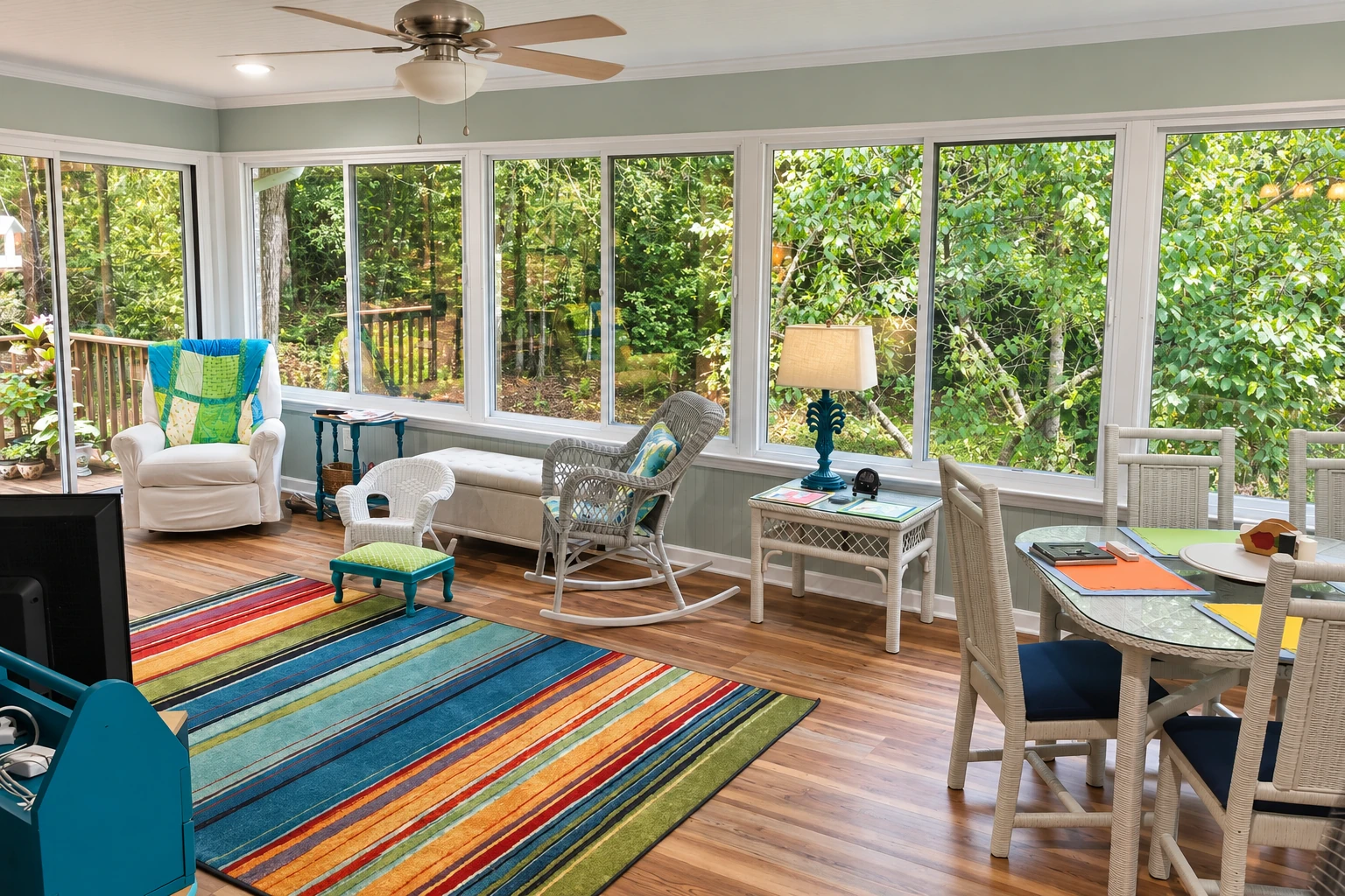 Converted deck sunroom with broad windows, wood flooring, a colorful rug and seating for dining and lounging.
