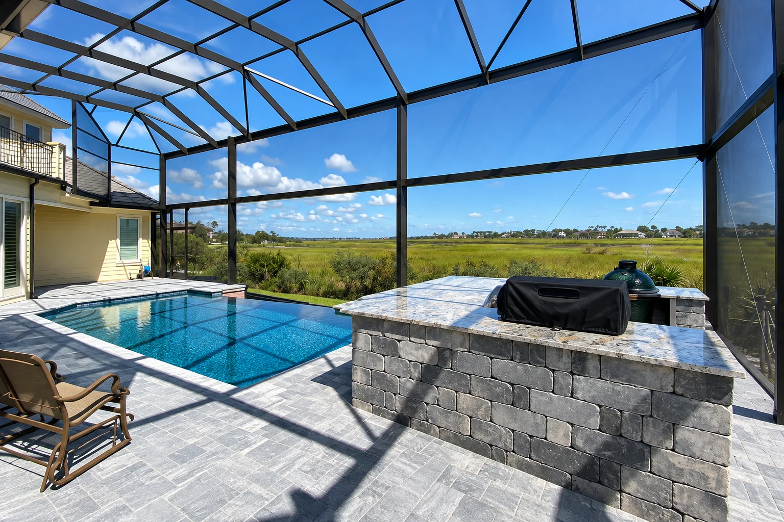 Enclosed patio with a black-framed screen roof, swimming pool, and stone outdoor kitchen overlooking a grassy marsh.