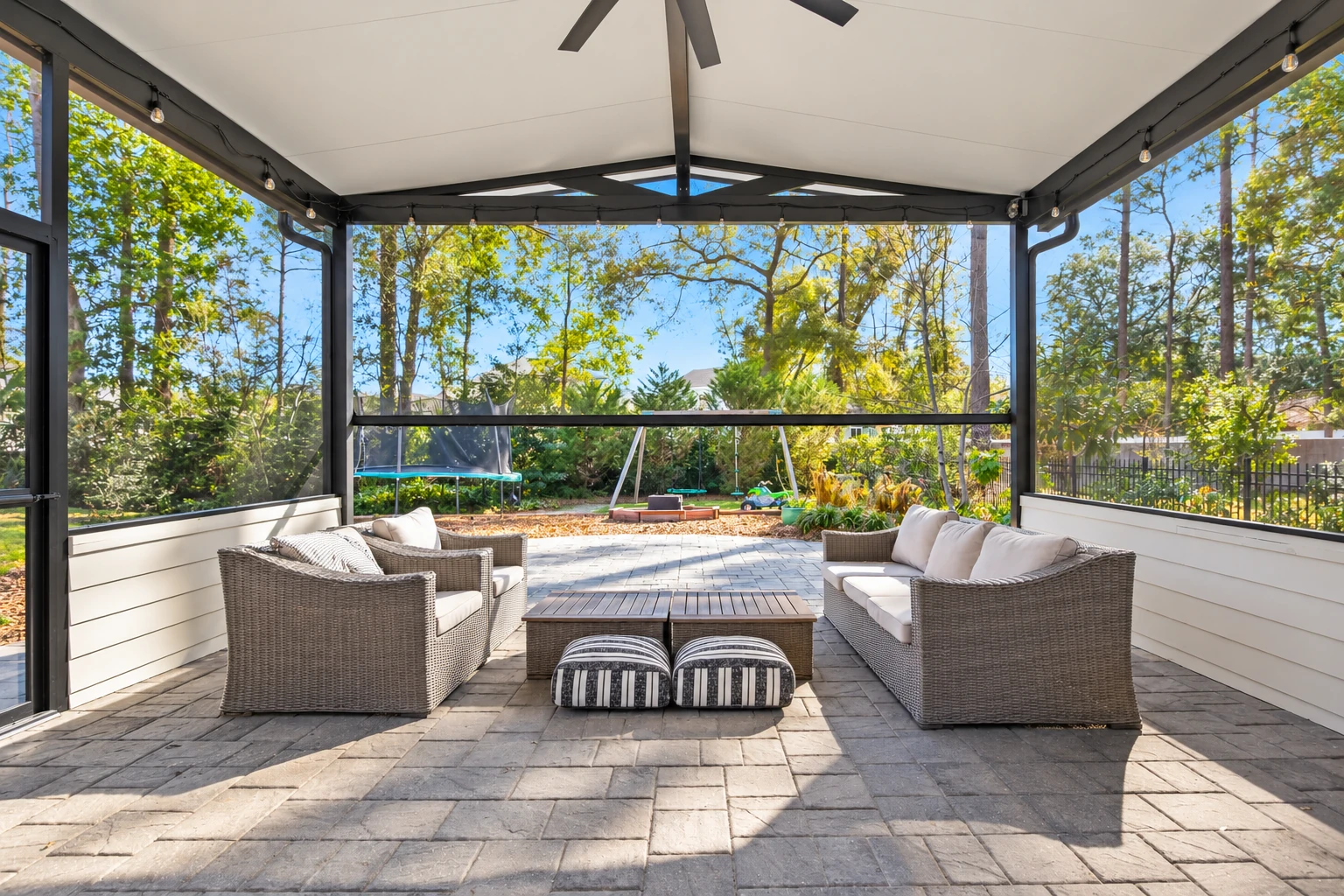A covered patio with a white ceiling, dark framing, and string lights shelters wicker sofas on a paved terrace.