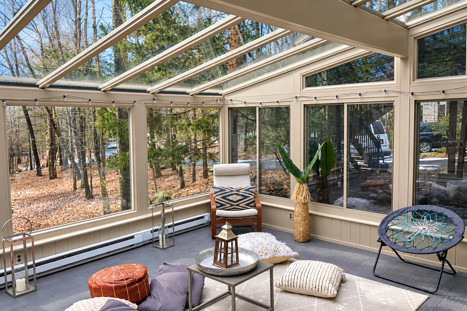 Bright solarium with a glass-paneled roof, large windows, string lights, and casual seating among wooded views.