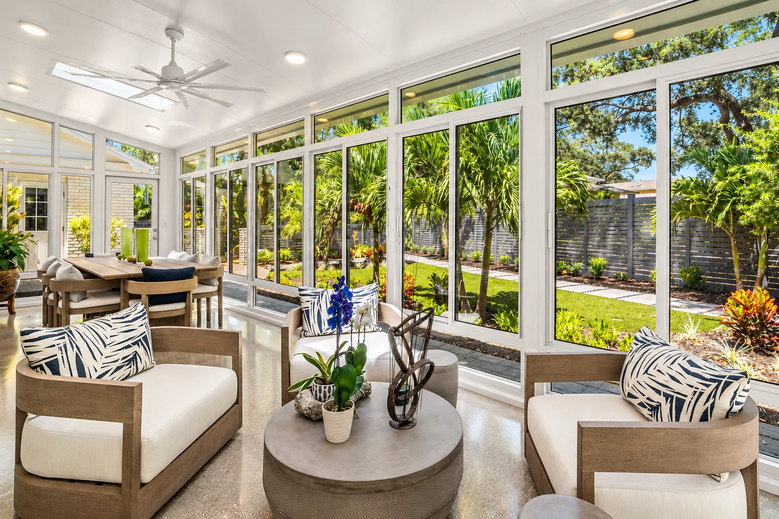 Bright remodeled sunroom with white-framed windows, neutral seating, and views of a lush garden.